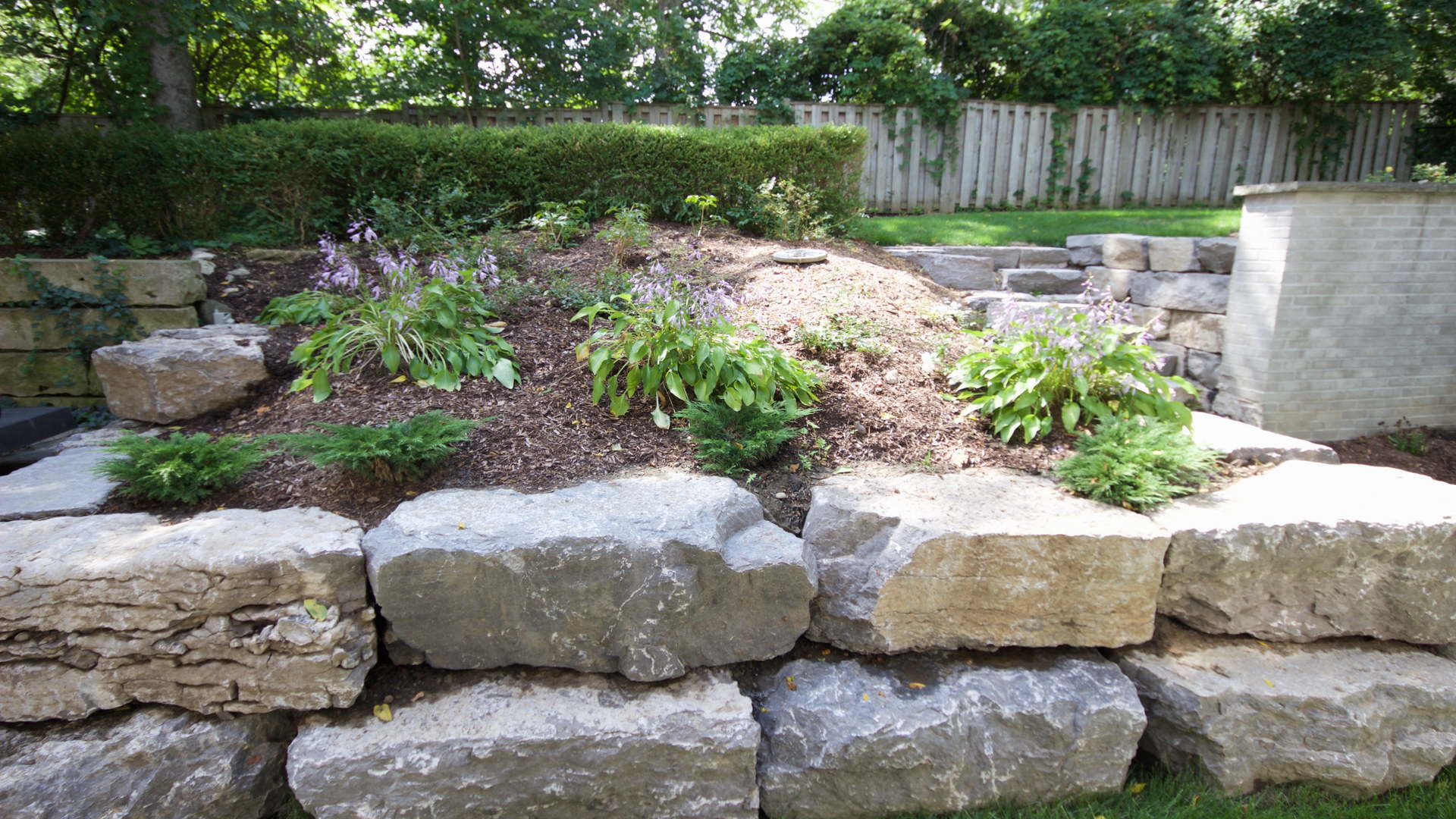 Natural garden with large armour stone retaining wall, various hostas and pine mulch. Traditional landscaping / hardscaping project in London Ontario.