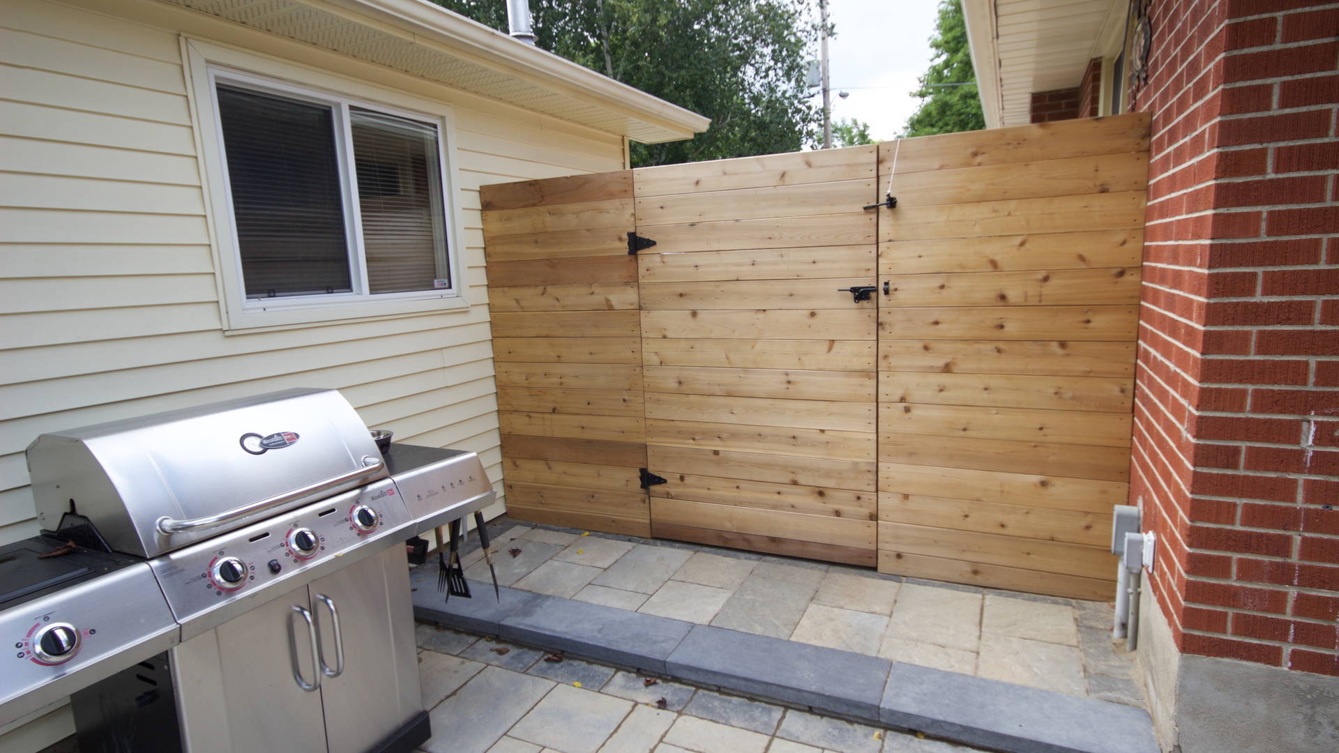 Small cooking area complete with a modern, horizontal fence and gate, and an interlock patio made up of large, pre-fabricated stones for the barbecue to rest on. Modern hardscaping and woodwork project in London Ontario.